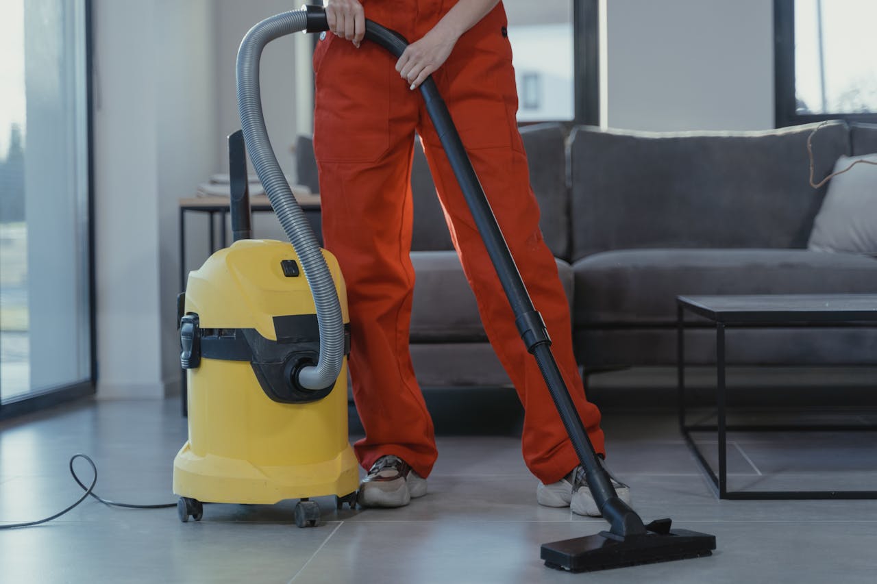 A professional cleaner in red coveralls vacuums a contemporary living room with a yellow vacuum cleaner.